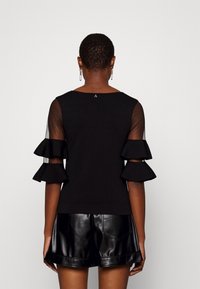 Schwarzes Oberteil mit transparenten, rüschierten Ärmeln und einer taillierten Passform. Kombiniert mit glänzenden schwarzen Leder-Shorts. Einfaches Design, minimale Akzente.