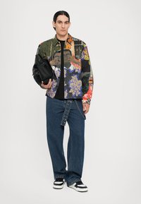 Mehrfarbig gemusterte Jacke mit floralen und grafischen Designs, schwarzes T-Shirt, breite Jeans und schwarz-weiße Sneakers.