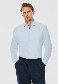 Jasnoniebieska koszula z długim rękawem z guzikami i kołnierzykiem. Miękka tekstura, dopasowany krój, połączona z ciemnymi spodniami z elastycznym paskiem.