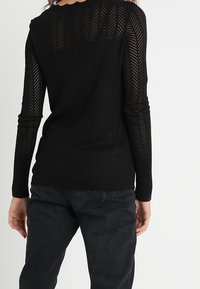 Kobieta ubrana w czarną, długą, dzianinową bluzkę z przezroczystym wzorem chevron na rękawach i górnej części pleców, w połączeniu z czarnymi spodniami, stoi tyłem.