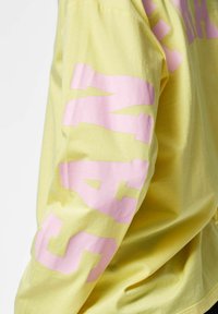 Żółta koszula z długim rękawem z dużymi różowymi literami "VANS" nadrukowanymi pionowo wzdłuż zewnętrznej strony rękawa.