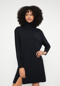 Vestido de malha com gola alta em azul-marinho escuro, mangas compridas, punhos e bainha canelados, com uma fenda lateral para detalhe adicional. Tecido macio e texturizado.