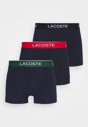 Tri pary mornarskih bokseric z kontrastnimi pasovi v rdeči, zeleni in črni barvi, vsi s logotipom "LACOSTE" in belimi poudarki.