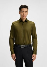 Olijfgroene blouse met lange mouwen, een getailleerd ontwerp, een puntcollar en witte knopen. Draagt met een zwarte broek en een zwarte riem.