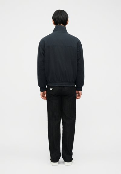 Άνδρας που στέκεται με την πλάτη γυρισμένη, φοράει μαύρο μπουφάν με ψηλό γιακά, μαύρο παντελόνι με άσπρη ετικέτα στην πίσω τσέπη και άσπρα παπούτσια.