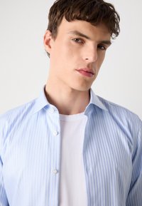 Camisa de vestir a rayas azul claro con una camiseta blanca debajo, que presenta cierres de botones y un collar clásico, textura suave y un ligero brillo.