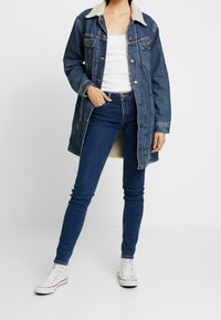 Jeansjacka med en pälsfodrad krage, blå färg, med knappstängning och två bröstfickor, bärs över en vit topp och blå jeans.