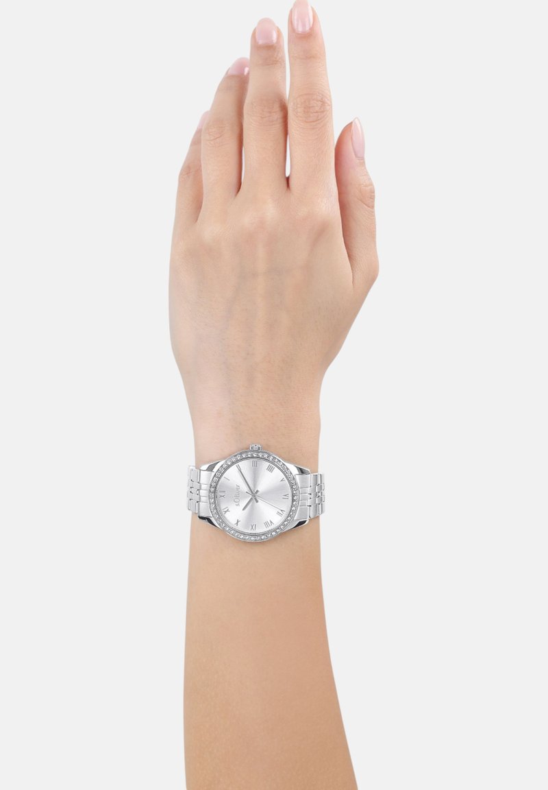 Silberne Armbanduhr mit rundem Zifferblatt, römischen Ziffern und einem mit Kristallen besetzten Gehäuse. Sie verfügt über ein Armband aus Metallgliedern und eine polierte Oberfläche.