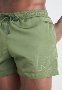 Olivenfargede badshorts med elastisk midjebånd og snor. Har stor "KARL"-logo i lys grå på venstre side. Myk tekstur.