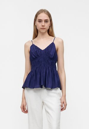 Jeune femme aux cheveux blonds et raides porte un haut bleu foncé sans manches froncé associé à un pantalon blanc ample, debout devant un fond blanc uni.