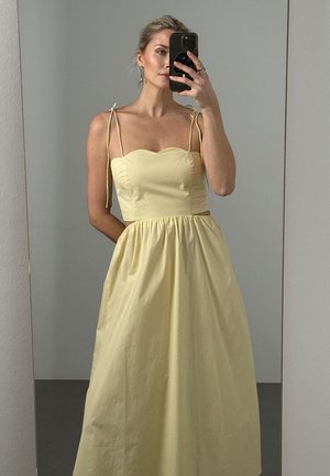 Femme prenant un selfie dans le miroir portant une robe sans manches jaune pâle avec de fines bretelles aux épaules et une jupe ample froncée.