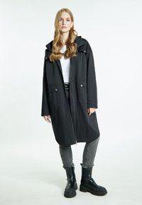Cappotto lungo nero con colletto, due tasche frontali e chiusura con zip. Abbinato a jeans grigi e stivali neri chunky. Tessuto liscio.