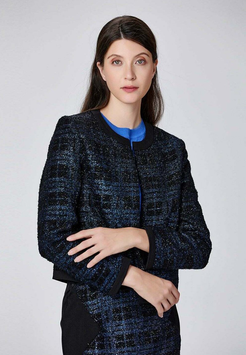 Veste texturée noire et bleue avec un col rond, des manches longues et une coupe droite, présentant de subtils détails scintillants et une garniture contrastante.