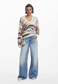 Gestrickter Pullover mit V-Ausschnitt und mehrfarbigen, wirbelnden Mustern; kombiniert mit hellblauen, weit geschnittenen Denim-Jeans und einer kleinen Umhängetasche.