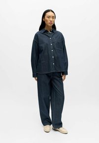 Donker denim overhemd met knopen en een zak op het borstvlak, gecombineerd met bijpassende broek met brede pijpen. Eenvoudig ontwerp met zichtbare stikseldetails.