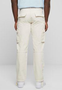 Pantalon cargo beige à coupe décontractée, avec deux poches sur les côtés, poches arrière à rabat et poignets élastiqués. Assorti à des baskets blanches.