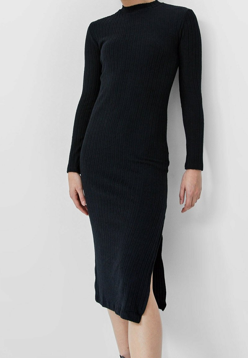 Femme portant une robe noire côtelée à manches longues, longueur midi, avec une fente latérale, debout devant un fond gris clair uni.