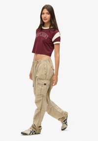 Afgesneden bordeauxrode t-shirt met witte mouwen met "Superdry" opdruk; gecombineerd met licht kaki cargo broek en stijlvolle sneakers.