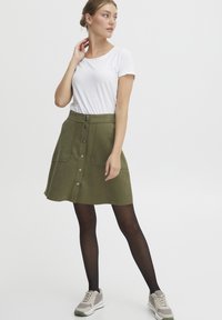 Olivenfarge A-linjeet skjørt med frontknapper, to sidelommer og en jevn tekstur; parret med en tettsittende hvit T-skjorte og sneakers.