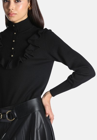 Femme portant un pull noir avec des volants et des boutons dorés, associé à une large ceinture en cuir noire et une jupe noire, vue de la taille vers le haut.