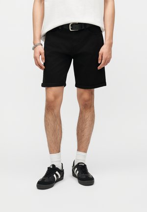 Mand iført optrukne sorte shorts, sort bælte, hvide crew-sokker, sorte og hvide sneakers, hvid skjorte og et sølvarmbånd, stående mod en hvid baggrund.