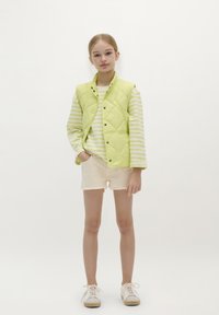 Chaleco acolchado verde lima sobre una camisa de rayas de manga larga, combinado con pantalones cortos beige y deportivas claras. Fondo blanco.