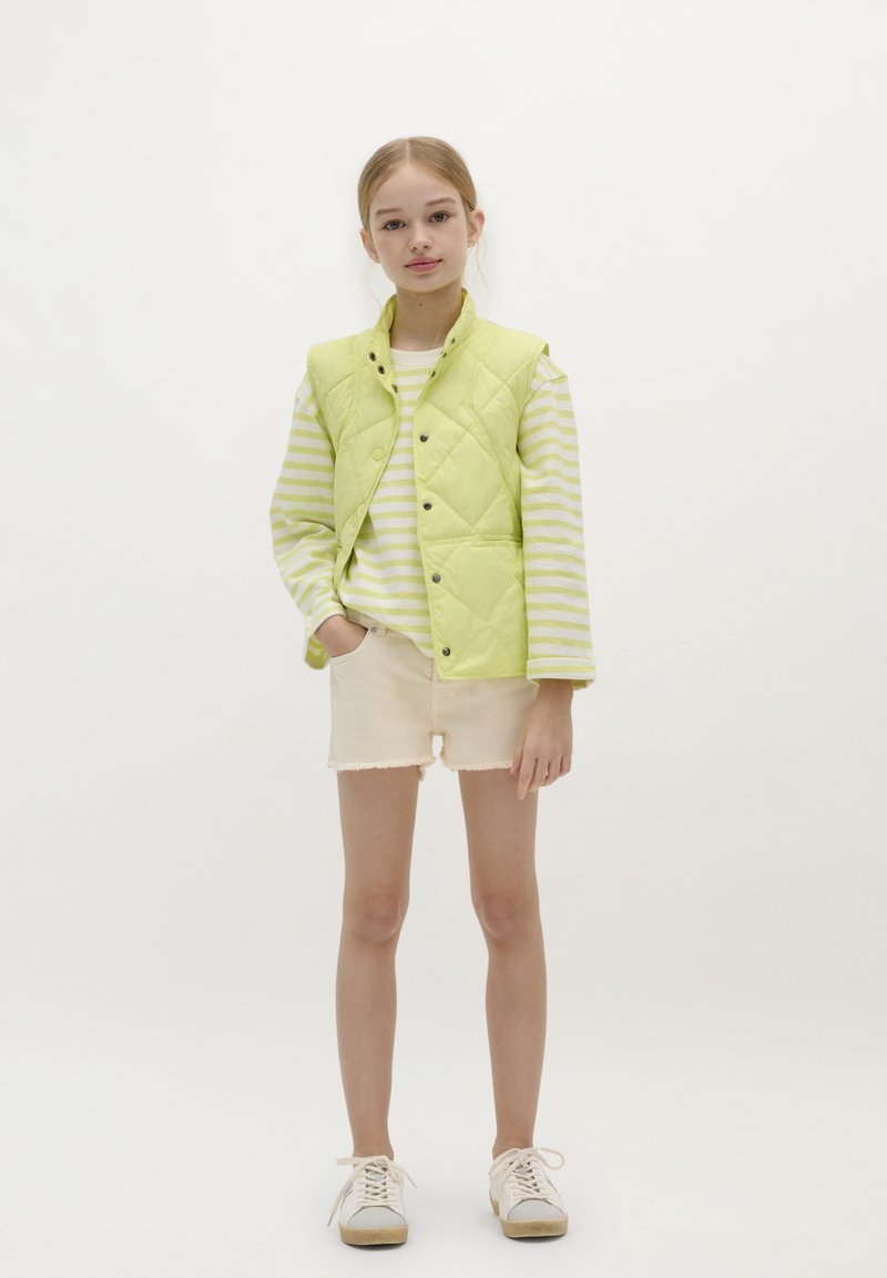 Chaleco acolchado verde lima sobre una camisa de rayas de manga larga, combinado con pantalones cortos beige y deportivas claras. Fondo blanco.
