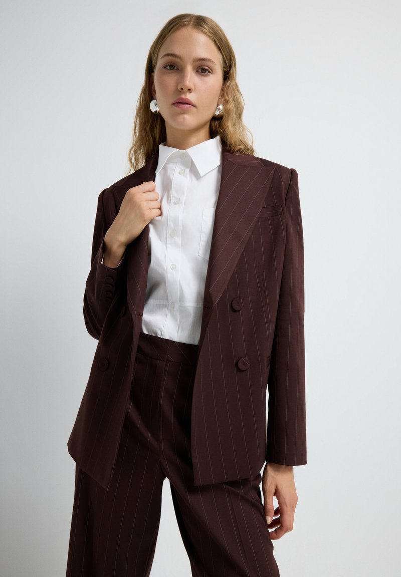 Mujer vestida con un traje de raya diplomática marrón oscuro, una camisa blanca de botones y pendientes de aro plateados, frente a un fondo gris claro.
