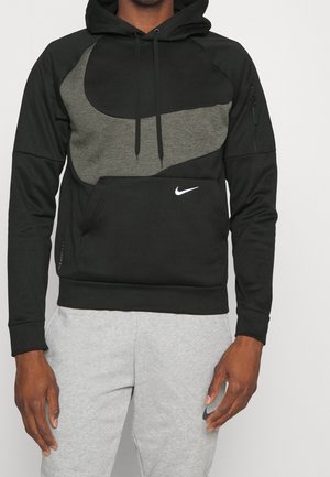 Homme portant un sweat à capuche Nike noir et gris avec poche kangourou et logo, associé à un pantalon de jogging gris clair avec logo Nike sur la cuisse gauche.