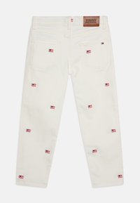 Pantalones de mezclilla blancos con pequeños diseños de banderas bordadas en azul, rojo y negro, y un parche de cuero marrón con el logo de Tommy Hilfiger en la parte trasera.