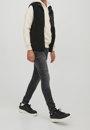 Schwarze Fleeceweste über einem cremefarbenen Hoodie, kombiniert mit dunklen Denim-Jeans und schwarzen Turnschuhen. Die Weste hat eine strukturierte Oberfläche mit Taschen.