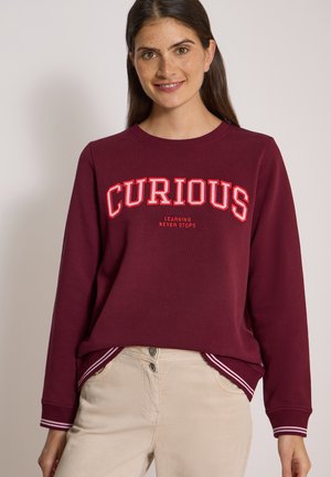 Femme portant un sweat-shirt bordeaux avec le texte "CURIOUS" et "LEARNING NEVER STOPS,", associé à un pantalon beige clair, souriant à la caméra.