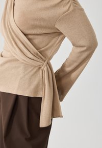 Beżowa bluzka o fasonie wrap, wykonana z miękkiego materiału, z drapowanym krojem, wiązaniem w talii i długimi rękawami, zestawiona z ciemnobrązowymi spodniami.