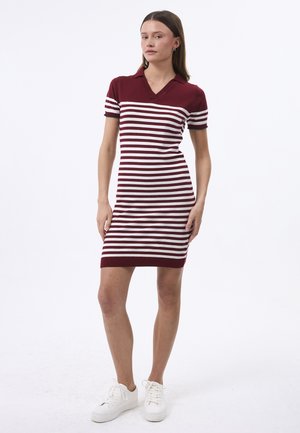 Kobieta stojąca w krótkiej sukience w poziome bordowe i białe paski z kołnierzykiem i białych sneakersach na białym tle.