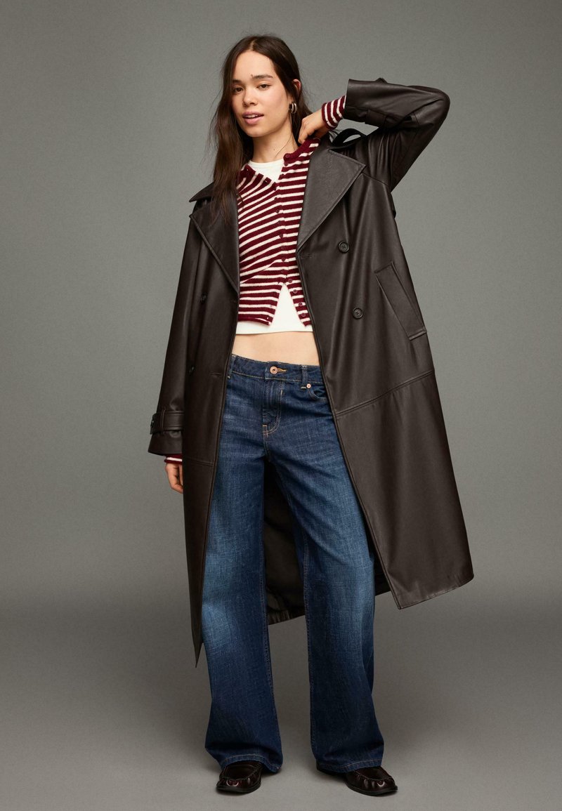 Długi czarny skórzany płaszcz typu trencz, krótkie sweter w paski w bordowo-białych kolorach, szerokie dżinsy o luźnych nogawkach, czarne buty oraz minimalistyczne detale w postaci akcesoriów.