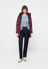 Bordeauxkleurige pufferjack met verticale stiksels, een blauwe cardigan over een grijze top, donkere spijkerbroek en lichtgrijze instap schoenen.