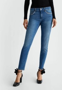 Mujer que lleva puestos jeans ajustados azules con bolsillos delanteros con tachuelas y zapatos de tacón negro con correas al tobillo y decoraciones florales.