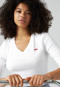 Bijela majica dugih rukava s V-izrezom i crvenim Levi's logotipom, s uklopljenim dizajnom i glatkim pamukom. Model drži kromirani rukohvat.