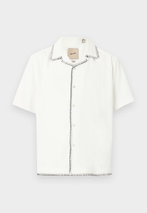 Chemise blanche à manches courtes avec boutons, détails de couture noire sur le col, la ligne de boutons et l'ourlet, présentée sur un fond clair.