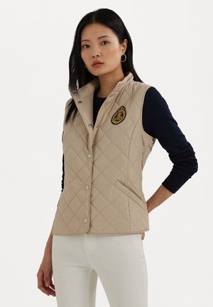 Gilet trapuntato beige con colletto rialzato, dotato di bottoni a pressione, tasche laterali e un dettaglio ricamato con uno stemma. Indossato sopra un top scuro.