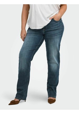 Kvinde iført mørkeblå jeans med lige ben, hvid top og brune spidse hæle, med den ene hånd i lommen og den anden hvilende ved siden af.
