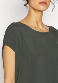 Top corto a maniche corte verde scuro realizzato in un tessuto morbido e liscio, con una ampia scollatura rotonda e una lucentezza discreta. Presenta un design minimalista.