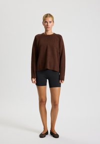 Hnedý pletený sveter s okrúhlym výstrihom a dlhými rukávmi, v kombinácii s čiernymi cyklistickými šortkami a čiernymi sieťovanými slip-on topánkami.