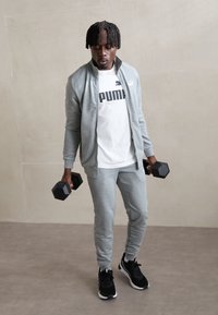 Ensemble de sport gris avec une veste zippée et un t-shirt à logo, tenant des haltères hexagonaux noirs, associé à des baskets noires.