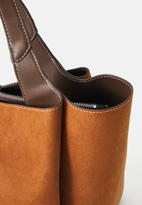 Bruine suede totebag met donkerbruine leren handvatten en zichtbare zilveren rits op een witte achtergrond.
