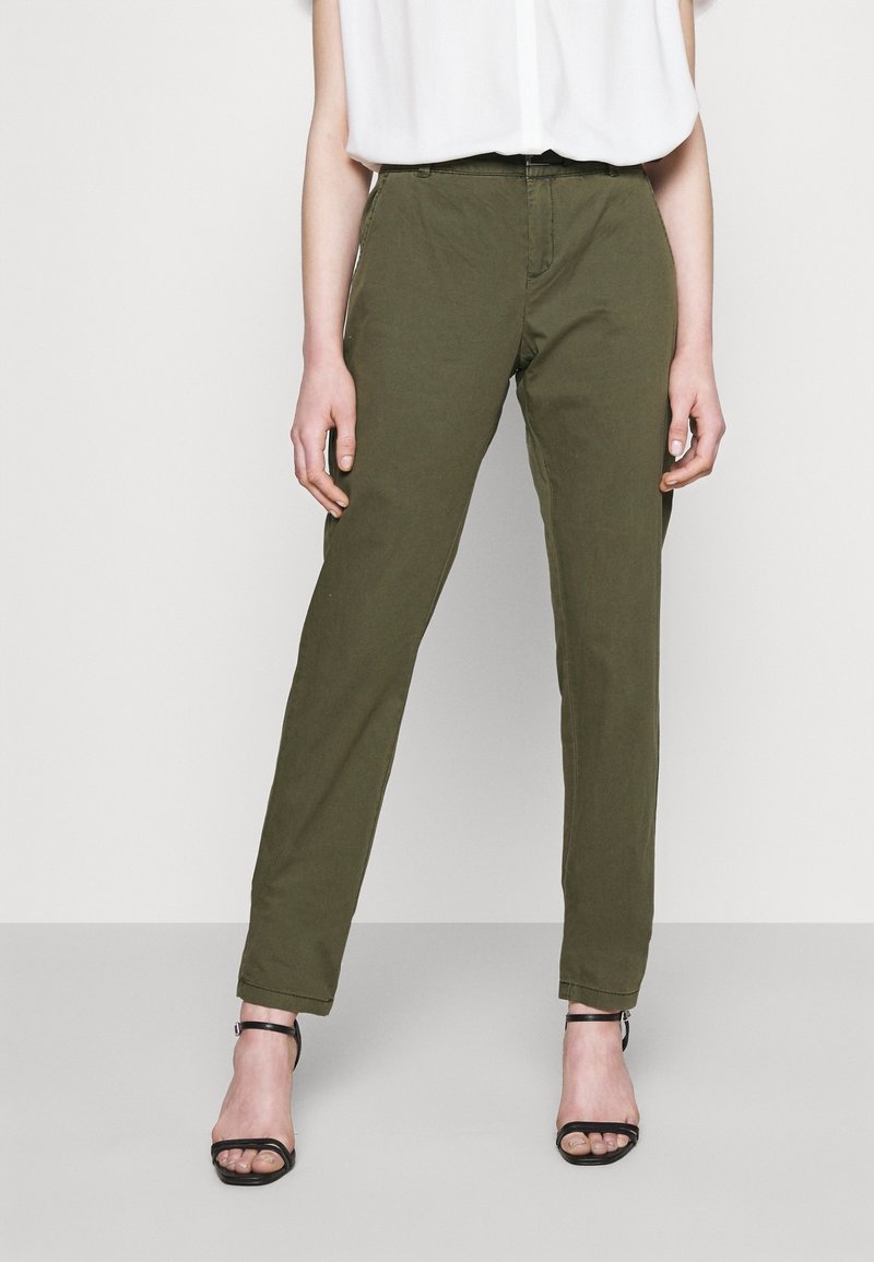 Femme portant un pantalon slim olive et des talons noirs à bride cheville ouverts à l'avant, debout devant un fond uni.