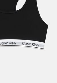 Czarny sportowy stanik Calvin Klein z białą elastyczną taśmą i czarnym logo na dolnej krawędzi, wyeksponowany na białym tle.