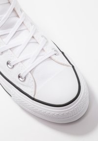 White canvas sneaker with white laces, metal eyelets, rubber toe cap, and black trim on a white background.