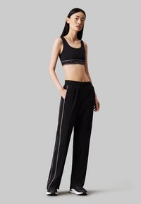 Crop top sportivo nero con logo Calvin Klein, abbinato a pantaloni neri con strisce laterali e cintura elastica. Il modello indossa scarpe sportive.
