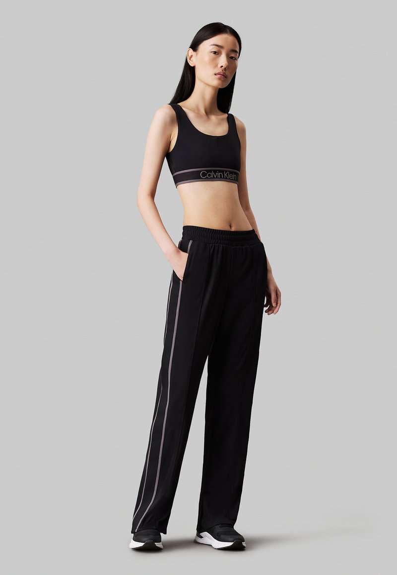 Crop top sportivo nero con logo Calvin Klein, abbinato a pantaloni neri con strisce laterali e cintura elastica. Il modello indossa scarpe sportive.